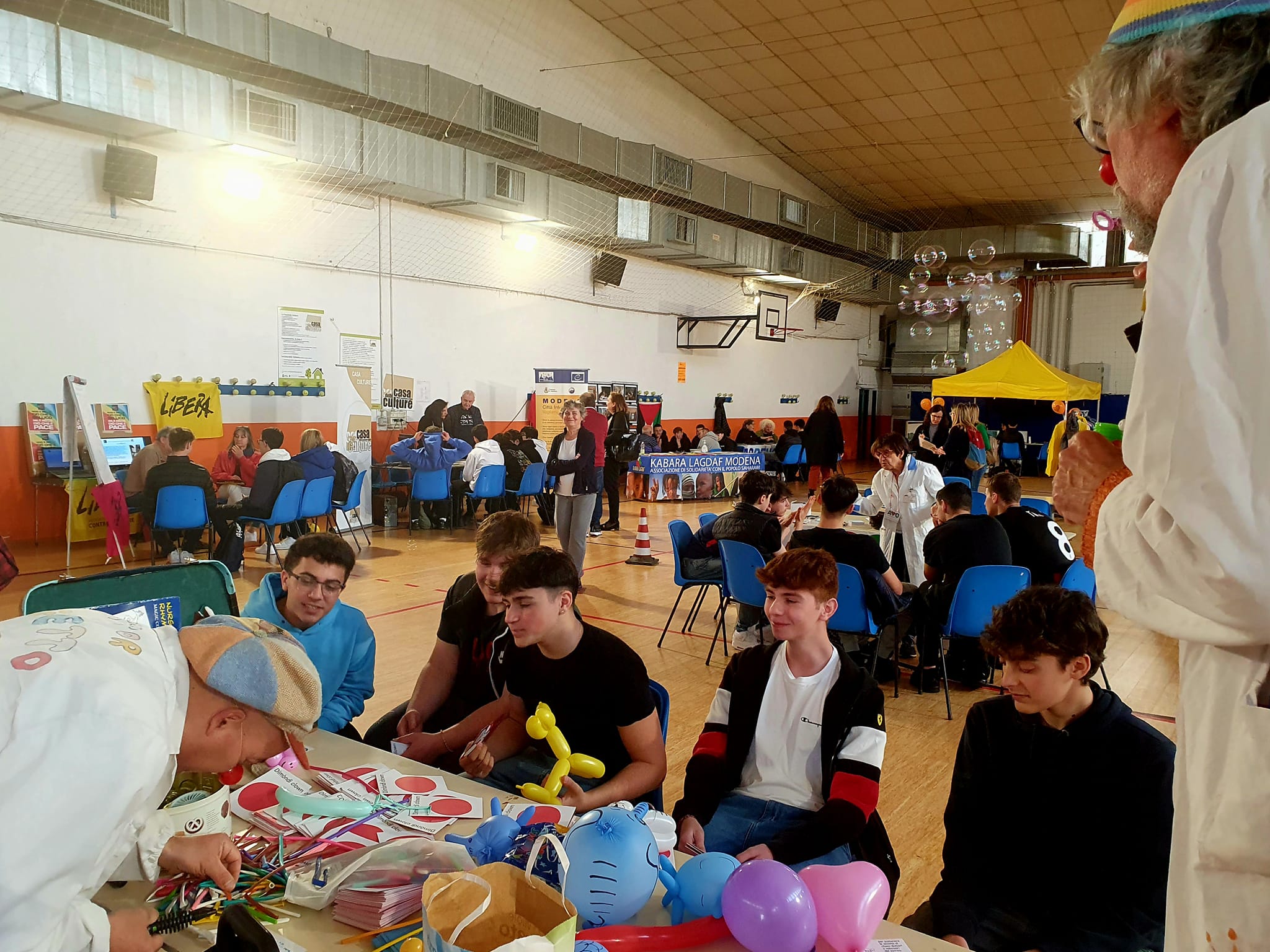 Un salone allestito con stand e un tavolo in primo piano circondato da ragazzi che osservano un uomo che svolge un laboratorio.