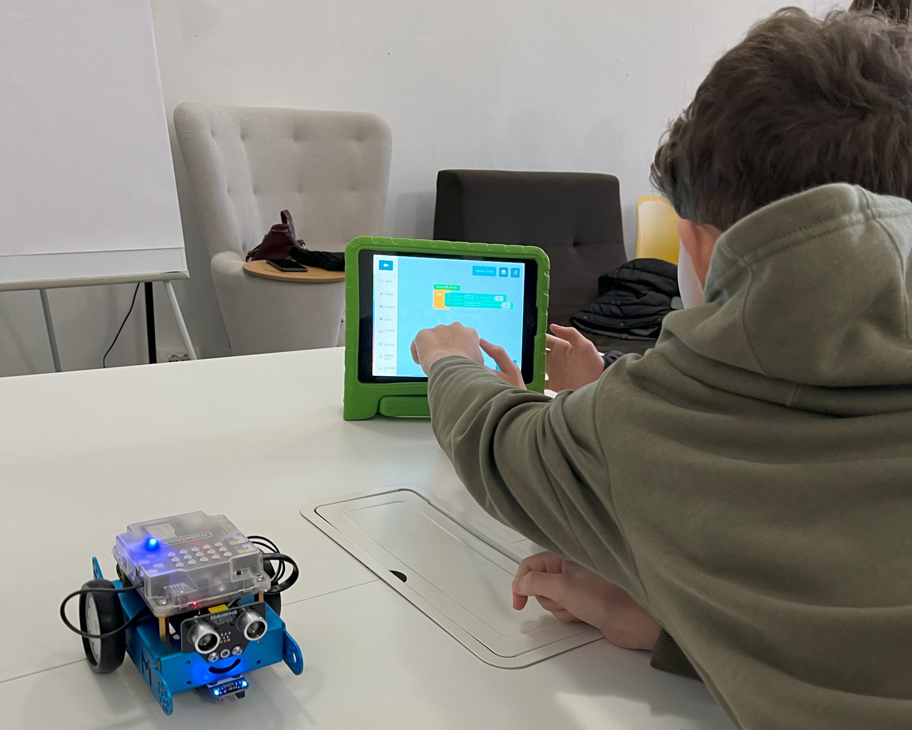 Un bambino usa un tablet avendo accanto un piccolo robottino per lavorare in classe.
