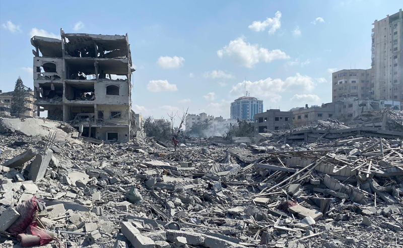 ampia distesa di macerie in primo piano e un edificio sulla sinistra con più piani parzialmente crollati e segni evidenti di esplosioni o incendi