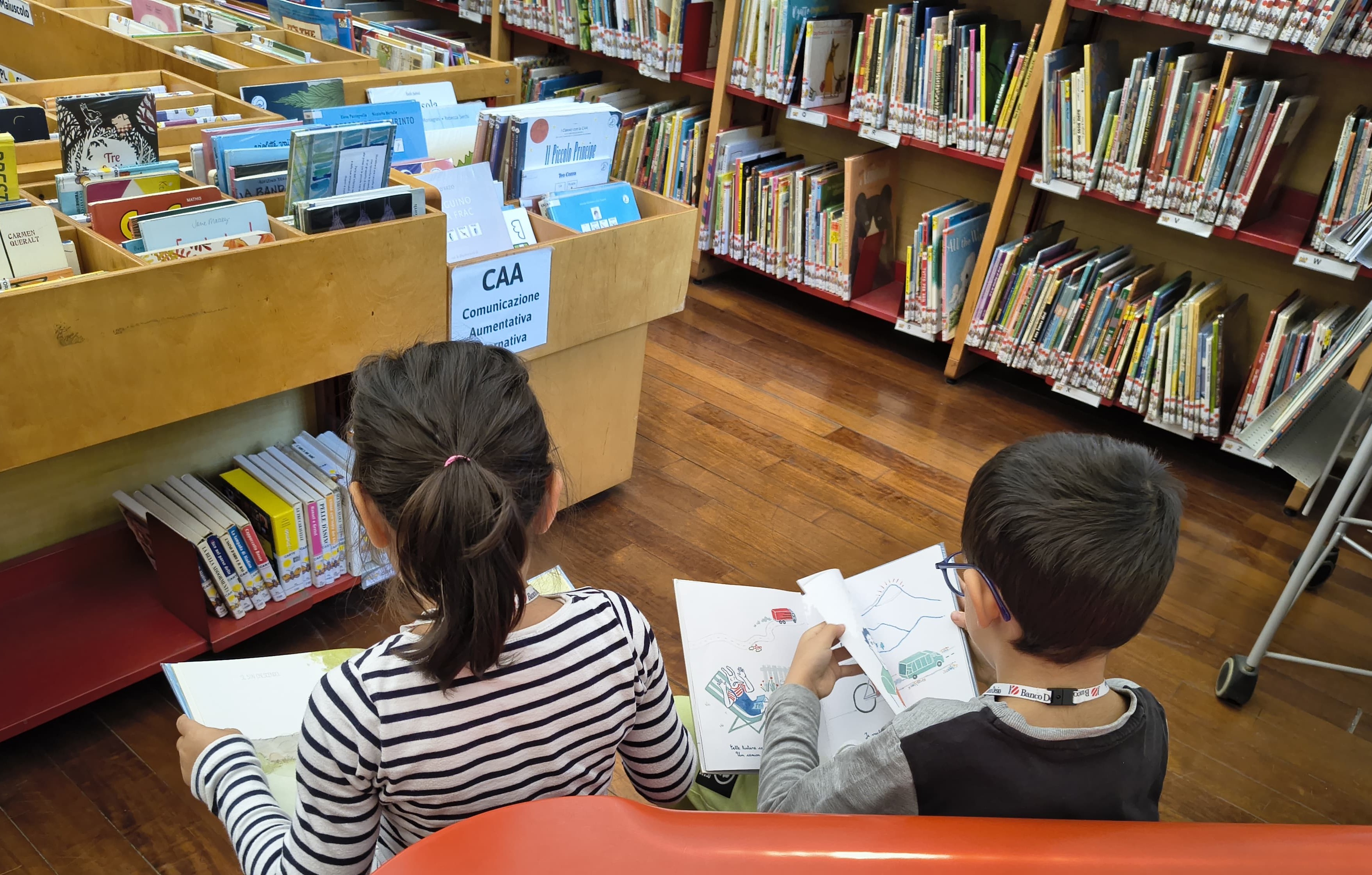 Due bambini seduti, ripresi di spalle, leggono due libri, in uno spazio della biblioteca. 