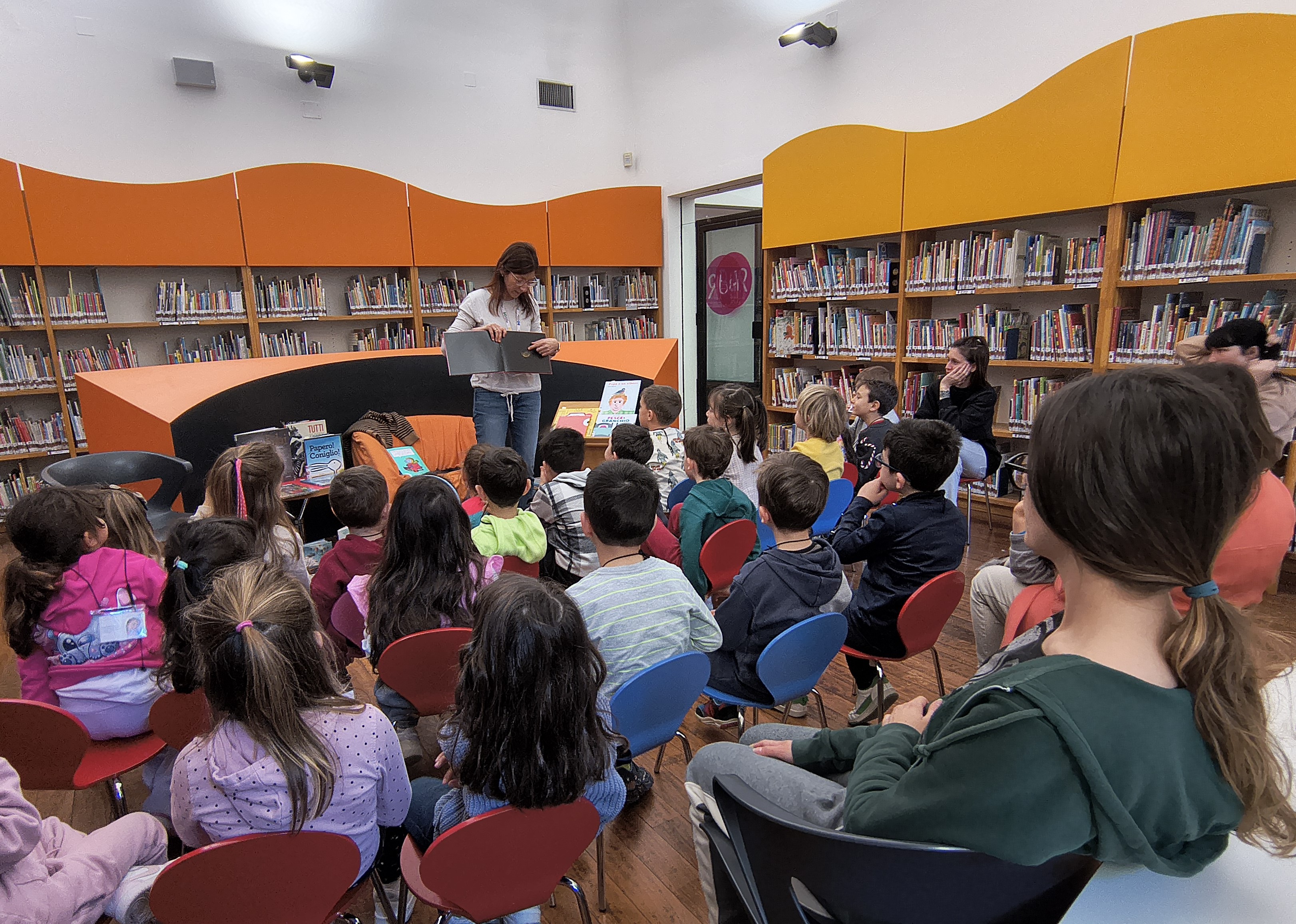 Una bibliotecaria sta mostrando le immagini di un libro a un gruppo di bambini, in una sala della biblioteca Delfini.