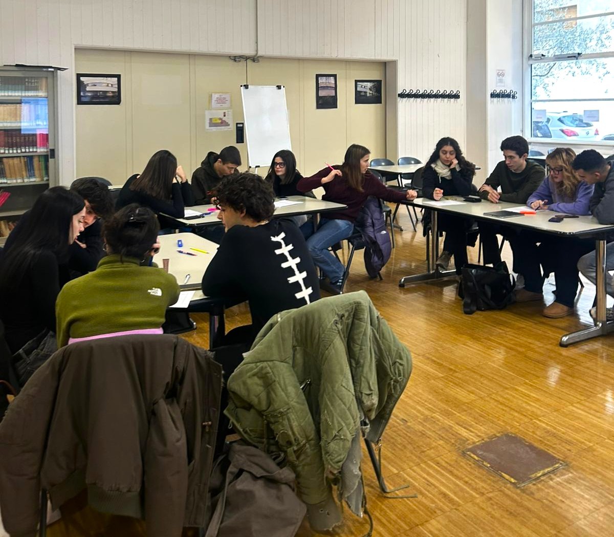 Un gruppo di studenti riunito in un’aula scolastica che lavora insieme attorno ai tavoli. Alcuni discutono, altri prendono appunti o svolgono attività