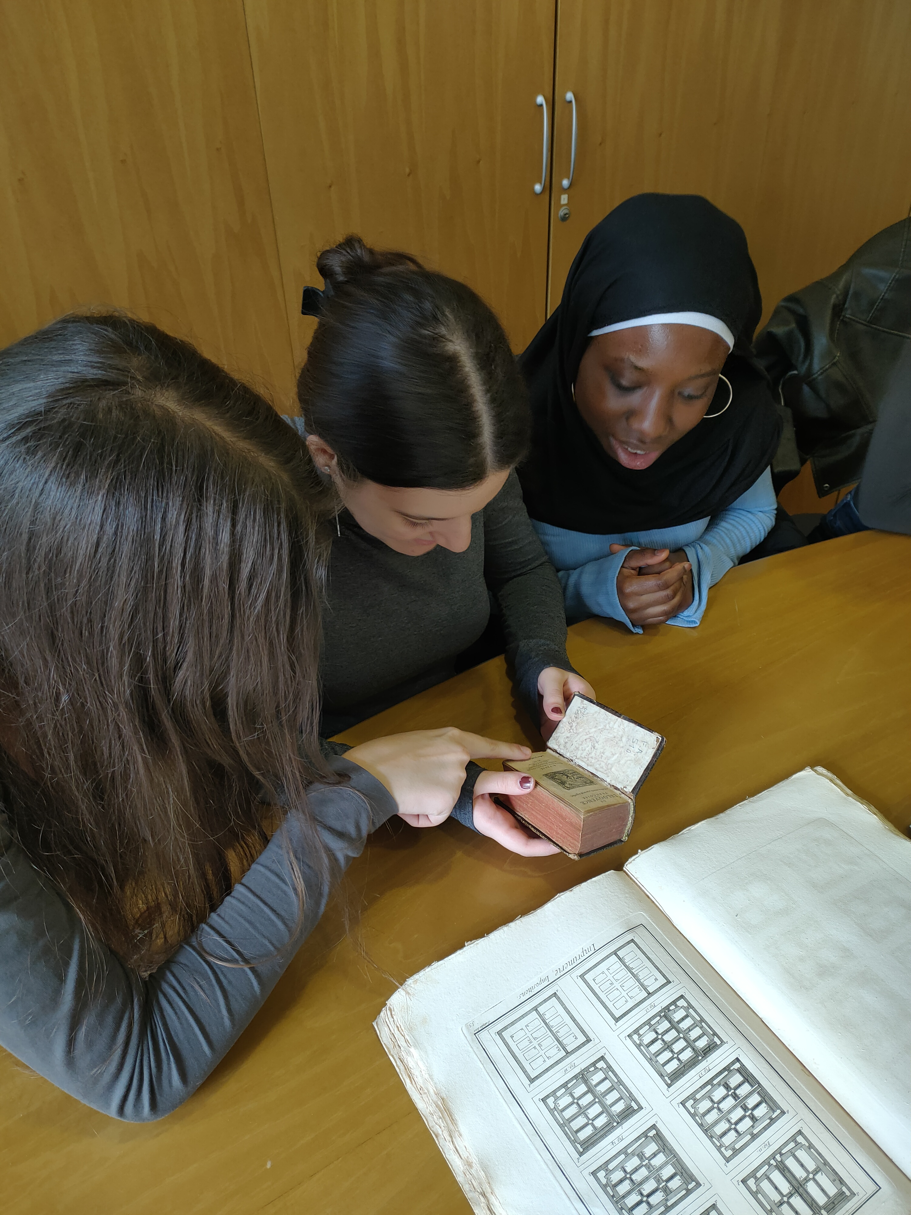 Tre ragazze osservano insieme alcuni libri antichi. Una di loro mostra una pagina, mentre sul tavolo sono aperti altri testi storici.