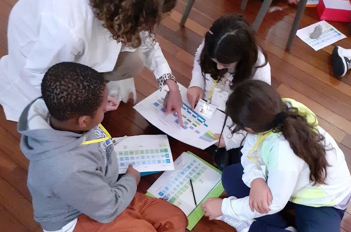 Un gruppo di studenti seduti a terra, lavora con l'esperta su tabelle scientifiche.