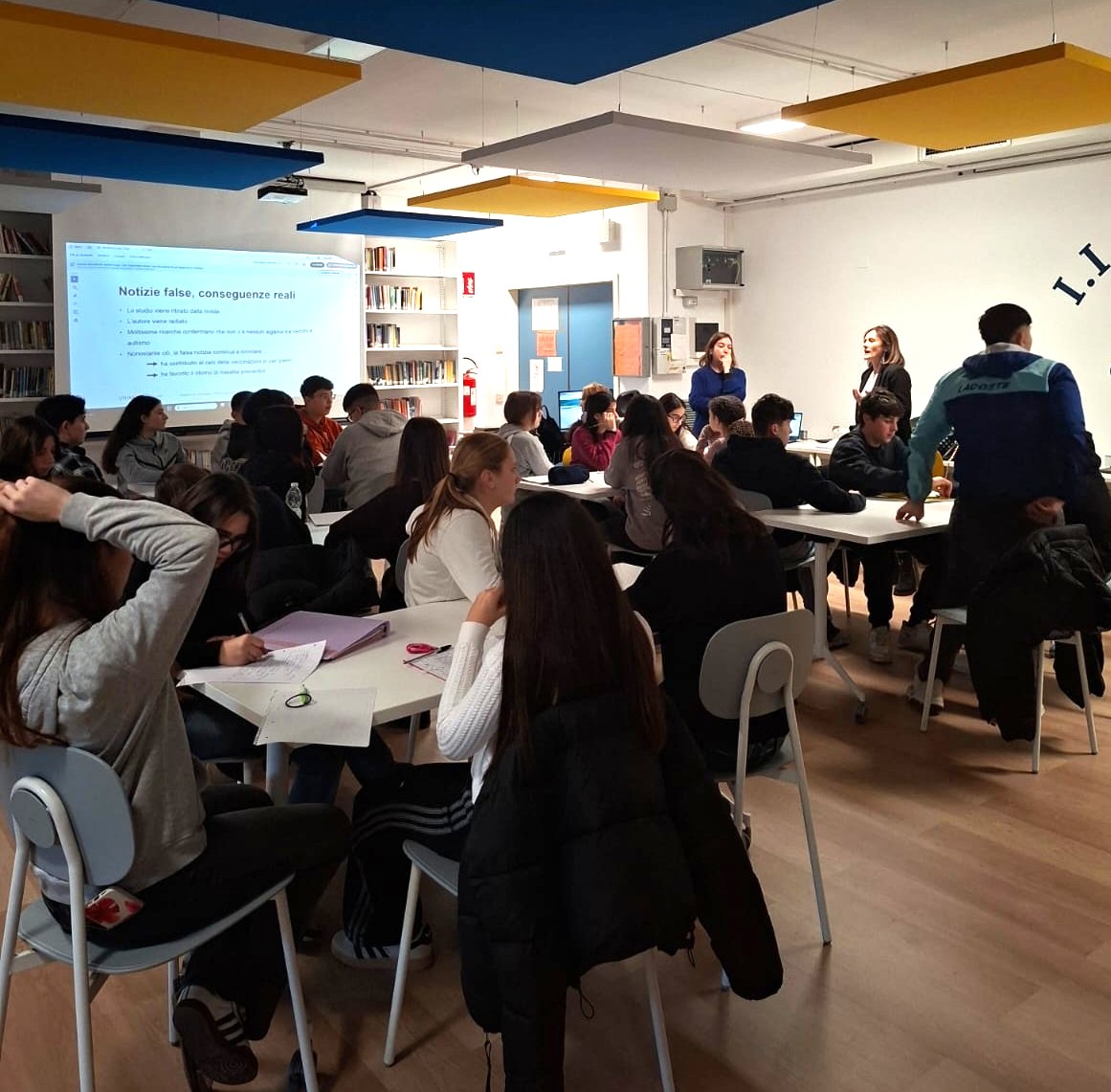 Gruppo di studenti che segue una lezione. Alcuni ascoltano gli insegnanti, mentre altri prendono appunti. Sullo schermo è proiettata una presentazione intitolata “Notizie false, conseguenze reali”.