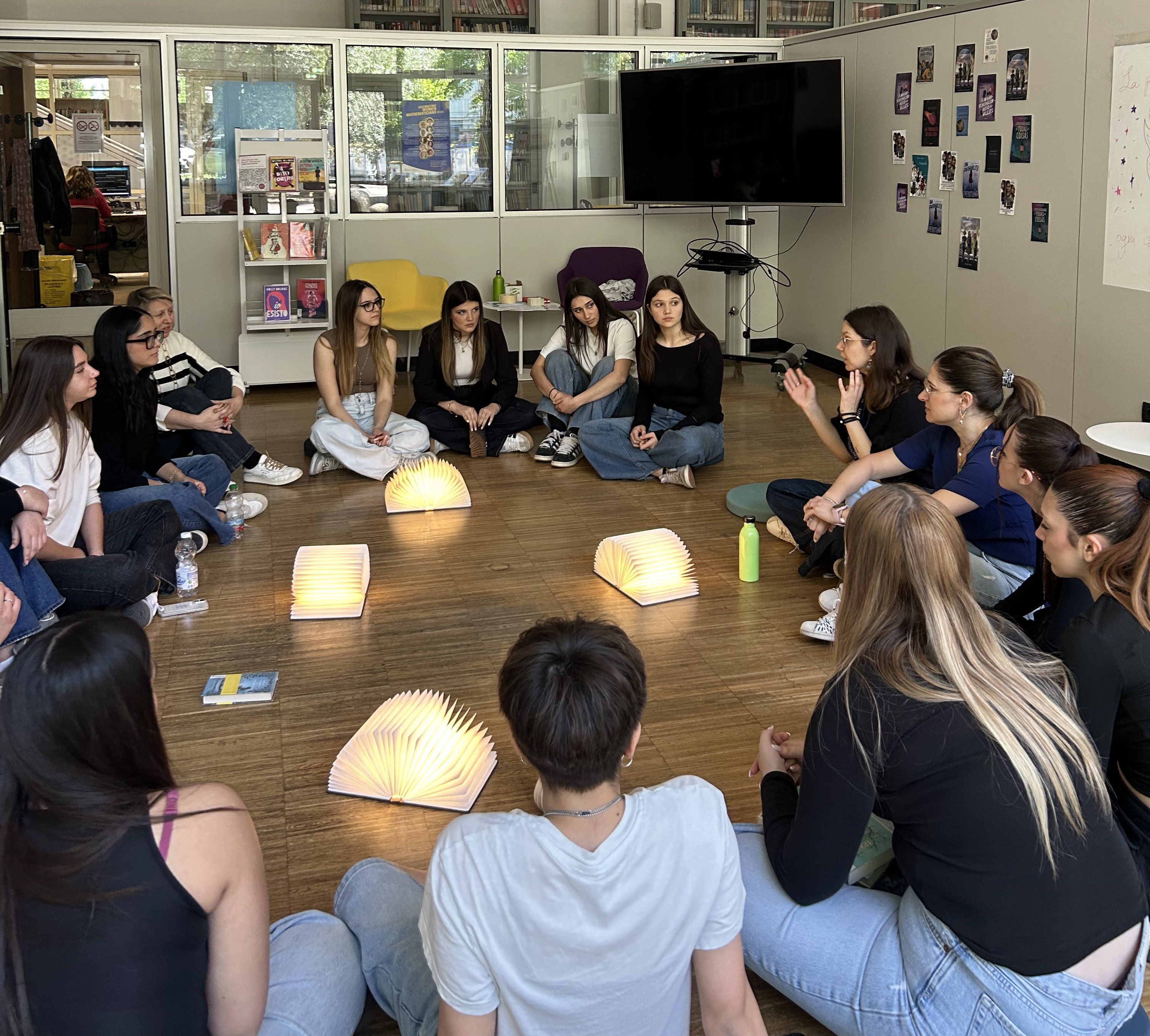 Un gruppo di ragazze e ragazzi seduto in cerchio in una sala, durante un momento di confronto. Al centro ci sono alcuni libri luminosi aperti