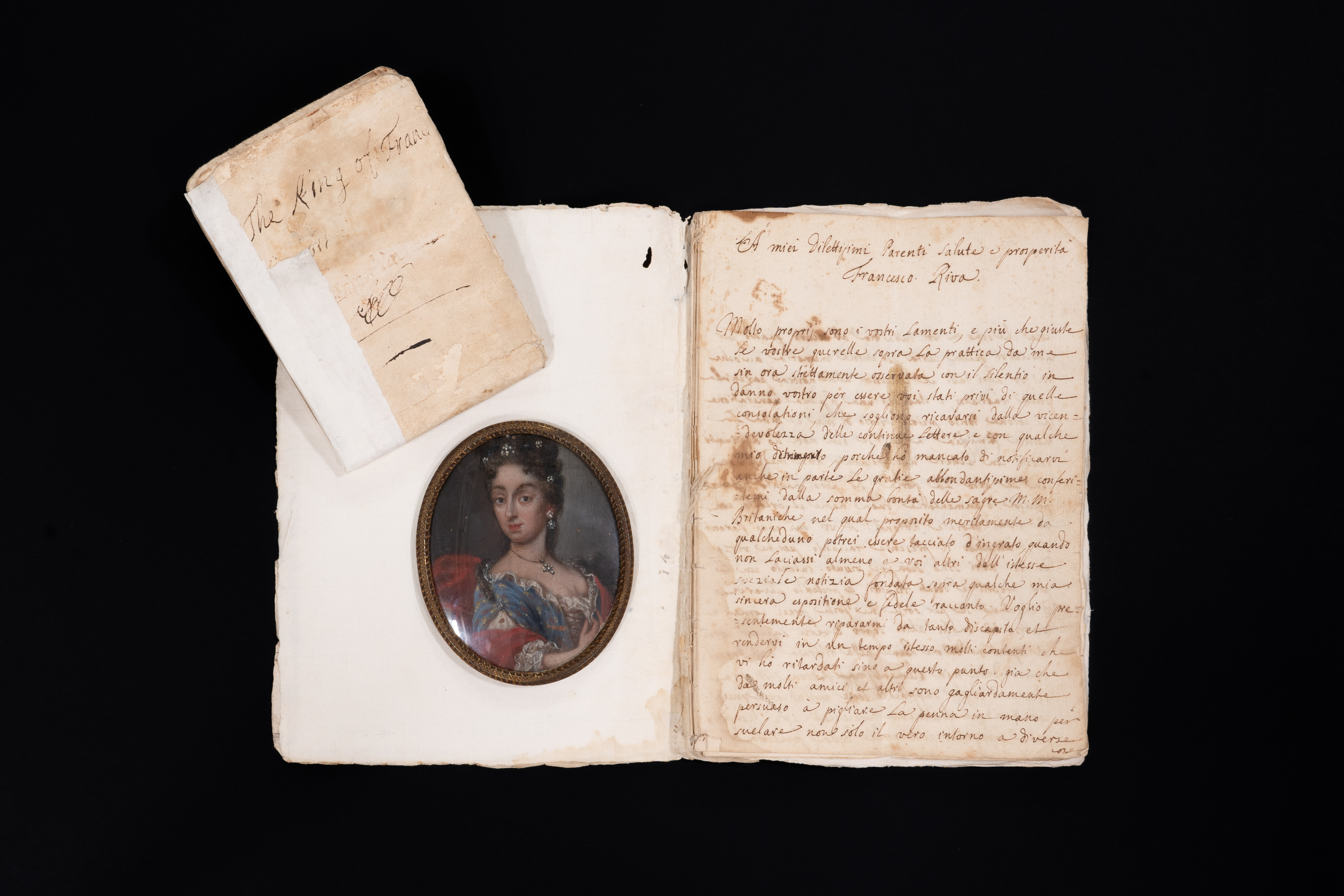Ritratto miniato di Maria Beatrice d’Este, raffigurata a mezzo busto con abito elegante e gioielli. La miniatura, inserita in un manoscritto seicentesco, è accompagnata da pagine manoscritte e da un piccolo fascicolo.